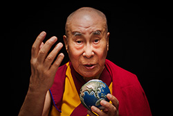 [asian man with shaved head, bushy eyebrows, maroon and gold robes, and red shawl draped on his left shoulder, speaks to camera while holding a small ball of the earth in with his left hand fingers, while gesturing with a raised right hand]