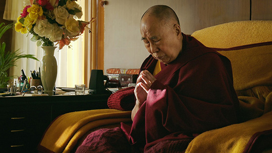 [asian man with shaved head, bushy eyebrows, maroon and gold robes, and deep red shawl draped over his shoulders, sits in a large chair covered in gold and maroon blankets to the left of a dark cabinet topped with a large bouquet of white, yellow, orange, and red flowers in a white vase, along with other personal items, with eyes closed and fingertips touching below his head tilted forward]