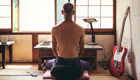 [a shirtless man with shaved head sits on a meditation cushion behind a low desk facing an open window with japanese screens, a red guitar leans against the wall at his side]